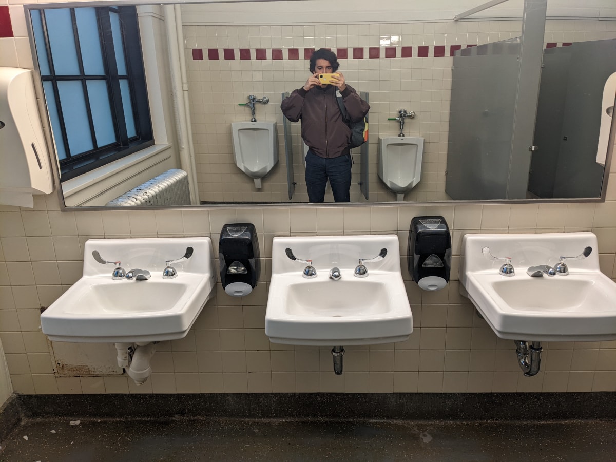 A selfie of Matthew taken in an MIT bathroom with three sinks beneath the mirror.