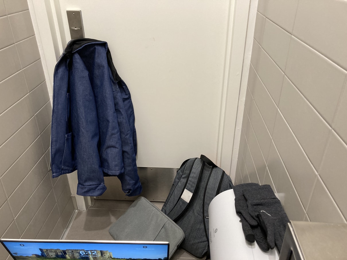 Backpack, laptop bag on floor of bathroom stall. Gloves resting on toilet paper dispenser. Jacket hanging up on handle of stall door. Laptop sitting on bare lap